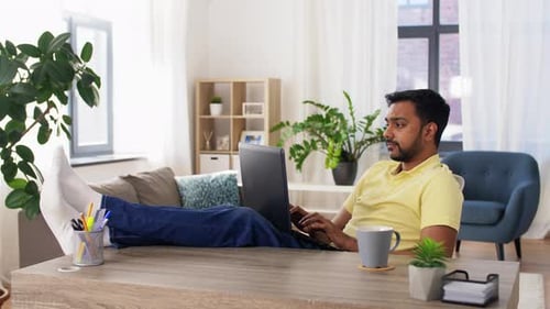 Tired young indian man working remotely with laptop at home office feet on table