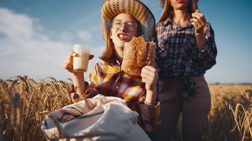 Young Girl Farmer in Sun Hat Eats Loaf of Bread Biting It Drinking Milk Sits on Wheat Field with
