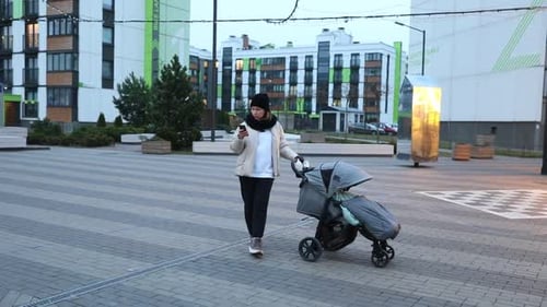 Woman with Stroller Checks Phone in Modern Urban Setting During Early Evening Hours