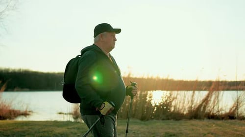 Active Handsome Elderly Man with Nordic Walking Sticks Walks Forward Through Along the Lake at