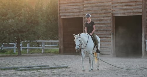 Young Woman Riding White Horse Training in the Paddock Near Wooden Stable at Sunset