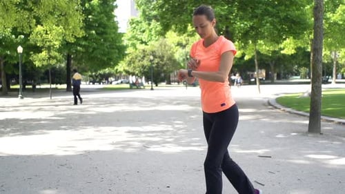 Young woman jogging and checking pulse on smartwatch in city park slow motion