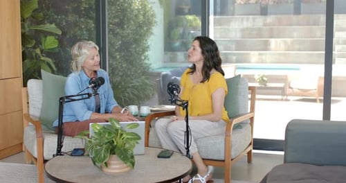 Two female hosts starting podcast in living room, left host adjusting mic, checking laptop