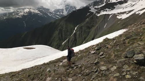 Aerial of Solo Hiker in High Mountains with Snowcapped Peaks Man Walks with Backpack Through Rocky
