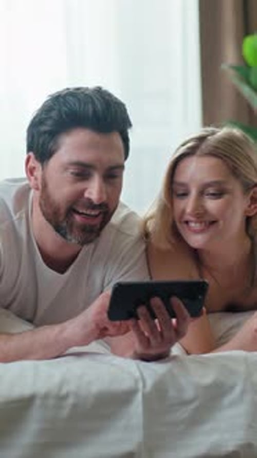 Alegre pareja caucásica usando el teléfono móvil riendo en la cama Familia viendo un divertido vídeo humorístico