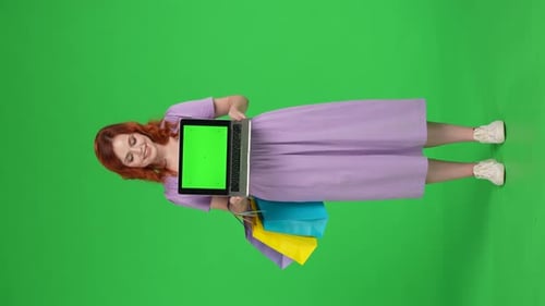 Redhaired Woman Holds a Laptop with a Green Screen Full Length Woman with Shopping Bags in Studio on