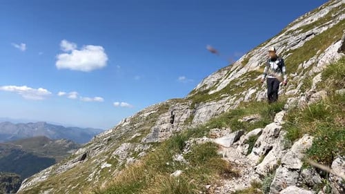 A Tourist with a Backpack and in Light Mountain Clothes on a Sunny Day Descends Along a Picturesque