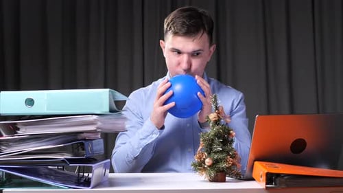 Office Worker Inflating Blue Balloon at Desk with Christmas Tree and Documents