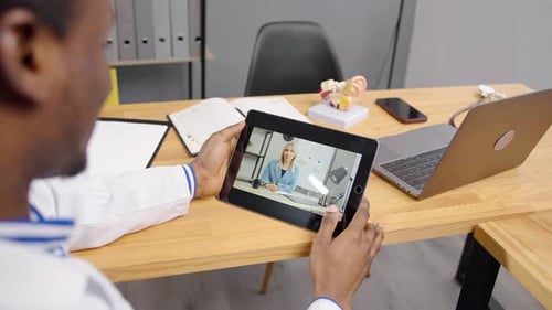 Black doctor sitting at office with tablet for an online consultation with female patient