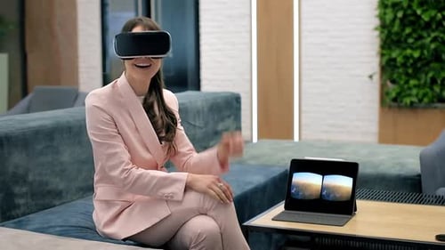A blonde caucasian excited woman exploring virtual reality using VR glasses in an office. Tablet on