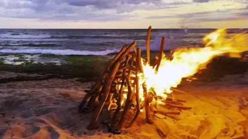 Lagerfeuer am Meer, am Strand, mit hellen, vom Wind verwehten Flammen und Meereshorizont in der Dämmerung