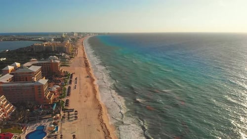 Drone flying towards tropical sea waves crashing on white sandy shore with a blue sky, golden sun ra