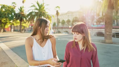 Two cheerful friends walking through the city while looking at their mobile phones and laughing