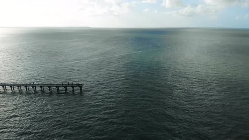 Ariel pullback of the end of a wooden pier that stretches into the vast, dark, gloomy, blue ocean on