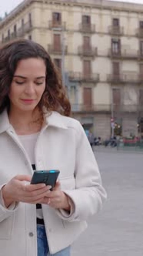 Cheerful Curly Young Woman in Casual Clothes Using Mobile Phone at City Street