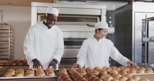 Portrait of happy diverse bakers working in bakery kitchen with fresh rolls in slow motion