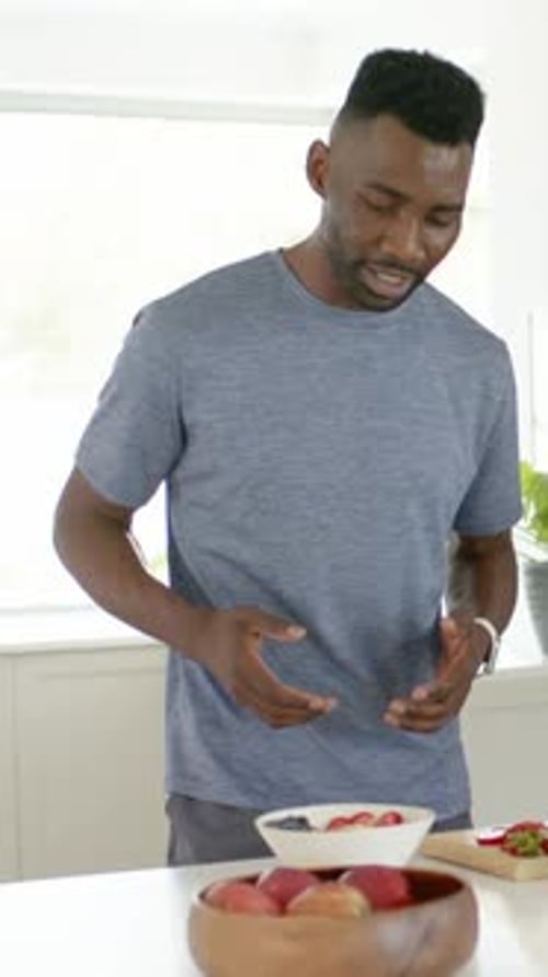Vertical video: Smiling man in gray tee gesturing placing white bowl at kitchen island to serve