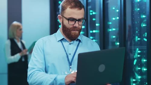 In a Server Room a Bearded Man Works Intently on His Laptop