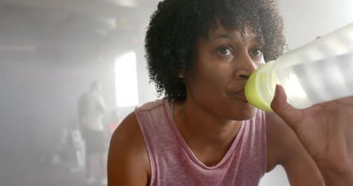 Tired african american woman resting and drinking water after training at gym, in slow motion