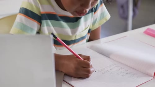 African American boy child doing math, writing in grid notebook with pink pencil at desk, striped