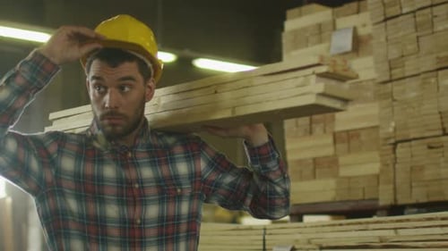 Lumber mill worker in hardhat is carrying wood in warehouse.