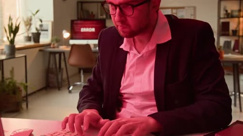 Irritated Man Removing Glasses Typing on Keyboard in Office with Red Light