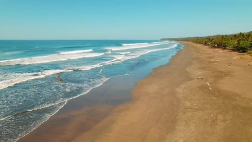 Aerial view of empty sandy beach with rolling waves under blue sky in Costa Rica