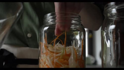 white housewife places carrots and cabbage for fermentation into a glass jar at home on the kitchen