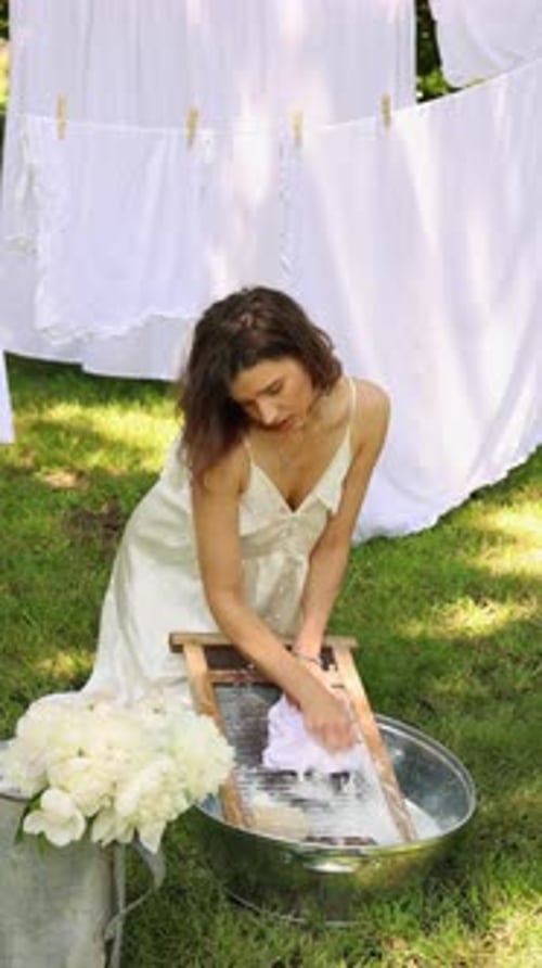 woman in dress is washing clothes on a washboard outdoors in a garden or park.