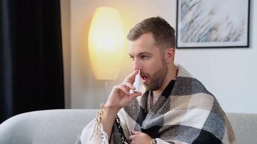Un jeune homme enveloppé dans une couverture vaporise un spray nasal assis sur un canapé