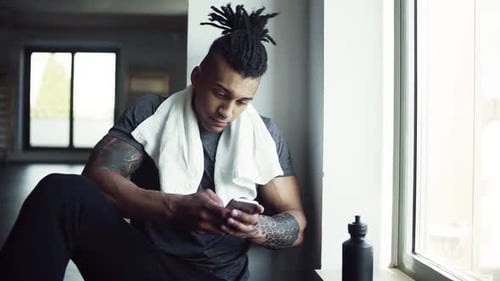 Young mixed race man texts on smartphone resting after workout at the gym