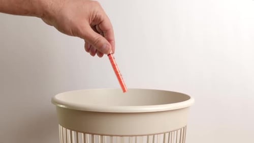 Hand Discards Orange Medical Syringe Into Trash Can