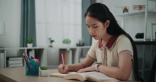Young asian woman sitting at desk writing information on diary notebook while reading book at home.