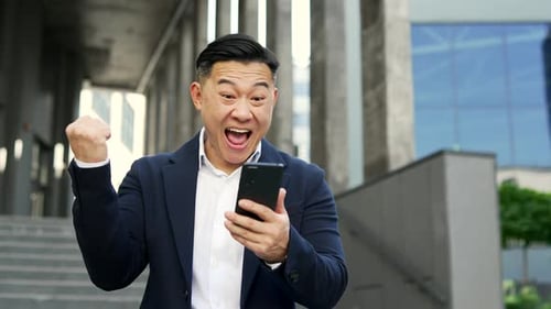 Asian businessman celebrates success after reading great news on smartphone while standing on street