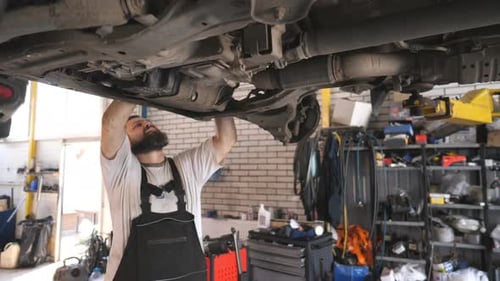 Professional Bearded Auto Mechanic Working Underneath a Lifting Vehicle at Service or Garage