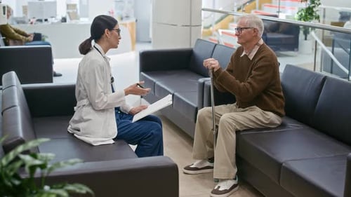 Elderly Man Communicating with Female Therapist Sitting in Hall of Modern Clinic