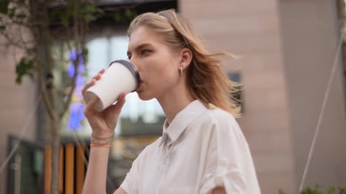 Side View of Pensive Caucasian Young Woman Sitting on Bench in City Street Drinking Coffee with