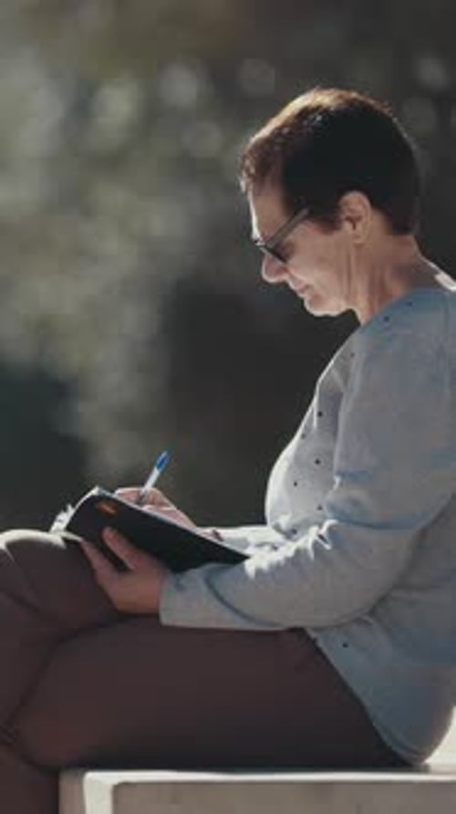 Woman Writing Notes in Notebook Sitting on Bench in Park