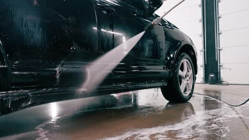 A male car wash employee applies car wash detergent to a black luxury car using spray gun