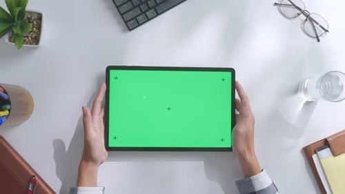 Top down view of hands holding a mock up green screen tablet on a white desk