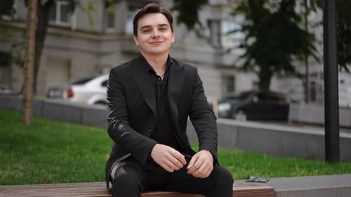 Un jeune homme en costume ajuste sa veste et sourit assis sur un banc dans un parc de la ville L'homme
