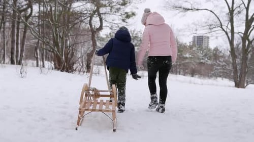 Cute Boy in Warm Clothing Walk with His Mother Pulling Wooden Vintage Sled on Snowy Winter Day