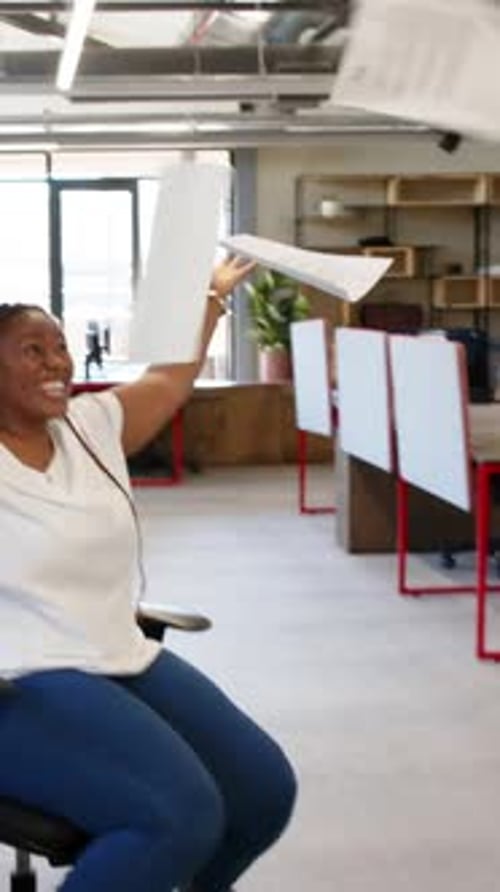 Vertical video: Diverse coworkers pushing and rolling chair across office after raising paper