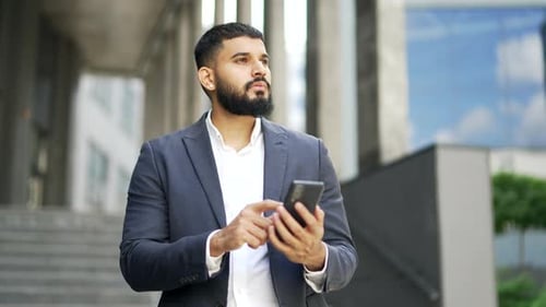 Thoughtful serious businessman is using a phone standing on street near an office building.