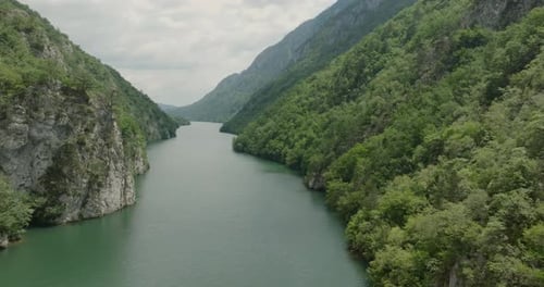 Drone flying forward above a beautiful river valley surrounded by steep cliffs and lush green woods