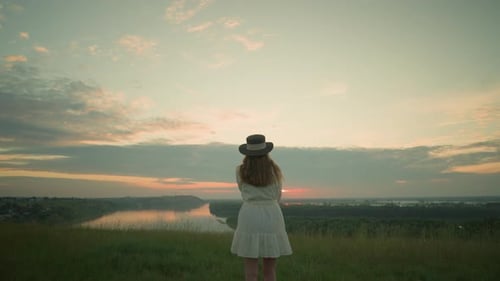 Woman in White Dress Gazing at Tranquil Lake During Serene Sunset in Grassy Field