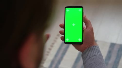 Young Man Sitting at Home Holding Smartphone Green Mockup Screen in Hand Male Person Using Chroma