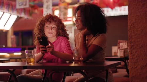 Two cheerful young women sitting together at a cafe table at night, with one friend showing somethin