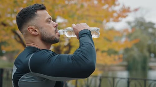 A Weary Male Athlete Soaked in Sweat Sips Water From a Sports Bottle After a Strenuous Jog in a City