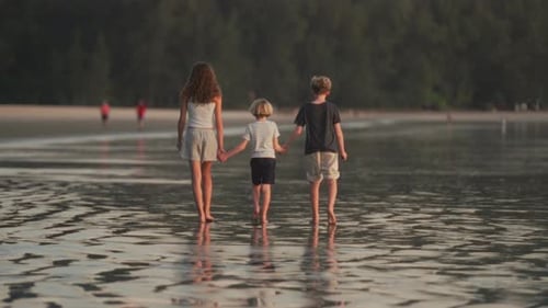 Siblings Walking Hand In Hand Along Shoreline With Lush Green Backdrop